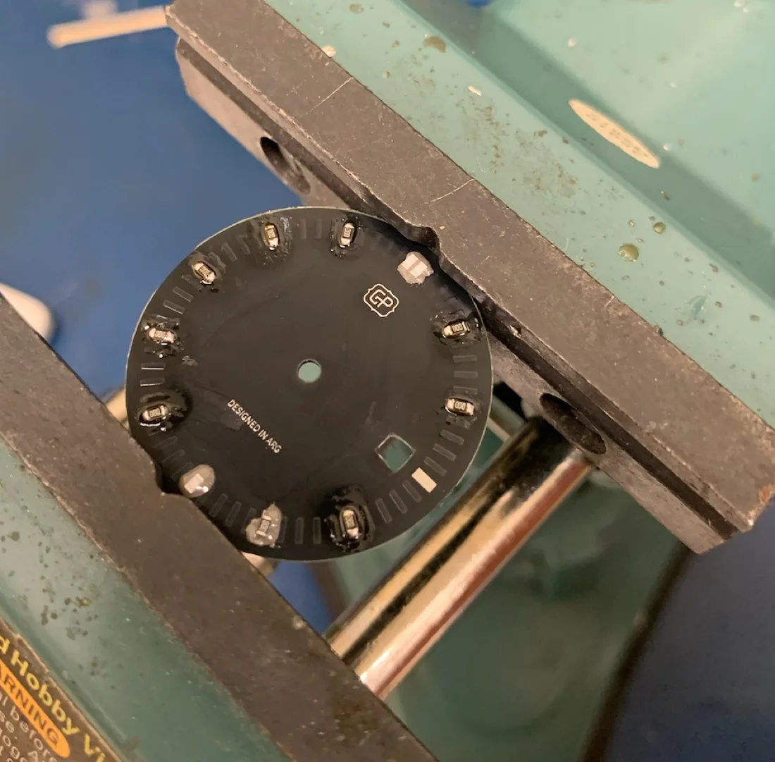 Custom black PCB watch dial held in a bench vise during soldering